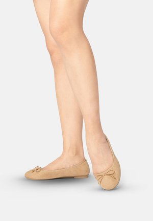 Frauenbeine, die beige Wildleder-Ballerinas mit kleinen Schleifen tragen, stehend auf einem hellgrauen Hintergrund.