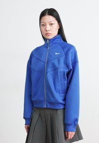 Blå Nike zip-up jakke med høy krage, med en tettsittende design, sidelommer og kontrasterende sømdetaljer, brukt over en grå plisserte skjørt.