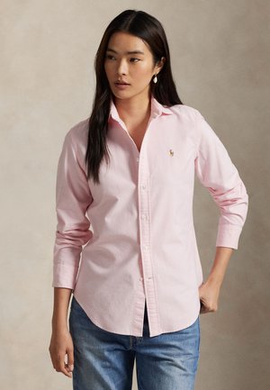 Kvinde iført en lys pink skjorte med knapper og blå jeans, stående med den ene hånd i lommen mod en ensfarvet baggrund.