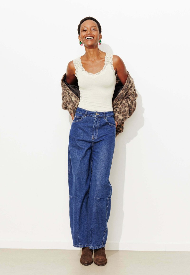 Débardeur blanc orné de dentelle, associé à un jean en denim bleu à jambes larges et taille haute. Bottines marron et châle imprimé léopard beige.