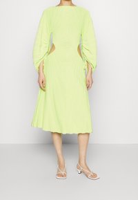 Abito midi plissé di un vivace verde lime, caratterizzato da maniche lunghe, aperture laterali e orlo svasato. Abbinato a sandali bianchi con fasce.