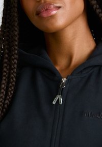 Hoodie preto com fecho de correr e acabamento texturizado, apresentando um fecho zip e um pequeno logotipo. O modelo tem cabelo trançado e lábios em tons neutros.