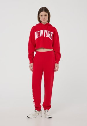 Czerwony krótki hoodie z białym napisem "NEW YORK", w połączeniu z dopasowanymi czerwonymi spodniami dresowymi z białym tekstem na nogawce. Białe sneakersy.