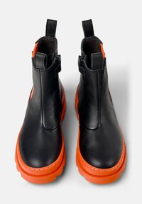 Bottines en cuir noir avec des semelles en caoutchouc orange, dotées d'une fermeture éclair sur le côté et de tirettes pour un enfilage facile. Texture lisse avec des accents contrastants.