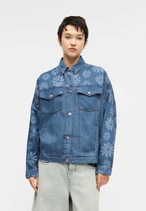 Personne portant une veste en denim bleu avec des motifs floraux clairs sur les manches et la poitrine, associée à un pantalon ample gris clair, debout devant un fond uni.