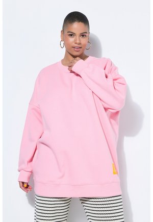 Femme portant un sweat-shirt rose oversize et un pantalon rayé noir et blanc, posant avec la main près du visage sur un fond blanc.