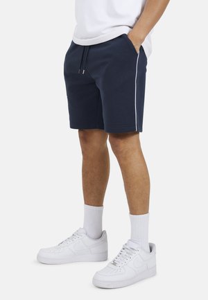 Mężczyzna ubrany w granatowe szorty z białym paskiem z boku, białe skarpetki za kostkę i białe tenisówki, stoi z rękami w kieszeniach.