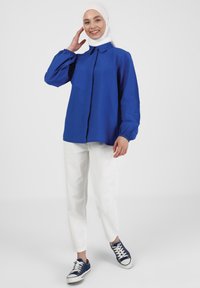 Camisa azul de botones con mangas acortadas, combinada con pantalones blancos y zapatillas de deporte marinas. La camisa presenta un cuello clásico y una textura suave.