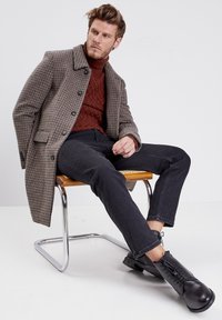Manteau marron à carreaux sur un col roulé en rouille texturé, associé à un jean noir et des bottes noires à lacets, assis sur une chaise en chrome.