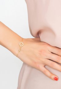 La pulsera de oro presenta un recorte floral adornado con piedras, unido a una delicada cadena. Textura suave con un acabado pulido.