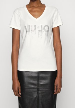 Person iført en hvid V-hals T-shirt med et pixeleret "LIU·JO" logo kombineret med en sort lædernederdel, vist fra nakke til hofter.