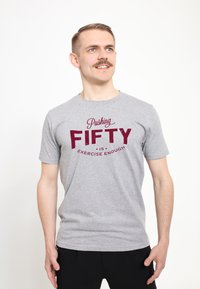 Šedé bavlněné tričko s tučným červeným nápisem "Pushing FIFTY is Exercise Enough." Krátké rukávy, kulatý výstřih, lehký materiál.