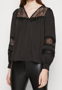 Svart blus med långa ärmar, spetsdetaljer längs halsringningen och ärmarna, samt en avslappnad passform. Slätt tyg, subtil plissering framtill.