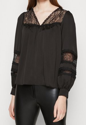 Blouse noire à manches longues, avec une bordure en dentelle le long du col et des manches, et une coupe décontractée. Tissu lisse, avec des plis subtils à l'avant.