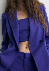 Blazer violet sur mesure à coupe structurée, doté de deux boutons noirs, associé à un top court assorti et un pantalon taille haute.