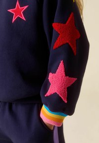 Tengerészkék pulóverujj texturált piros és rózsaszín csillagfoltokkal, színes csíkos mandzsettával, és kéz, mely a hozzá illő tengerészkék nadrág zsebében van.