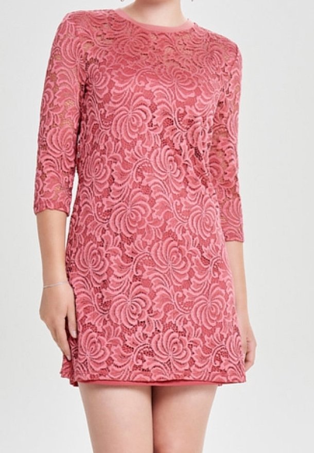 Femme portant une robe en dentelle rose à motifs floraux et manches trois-quarts, debout devant un fond clair uni.