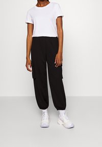 Pantalon cargo noir à coupe ample, avec des poignets élastiques et des poches latérales, porté avec un t-shirt blanc à manches courtes et des chaussures de sport blanches.