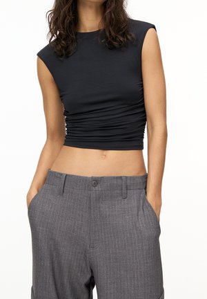 Femme portant un crop top noir sans manches froncé et un pantalon taille haute gris à fines rayures avec les mains dans les poches.