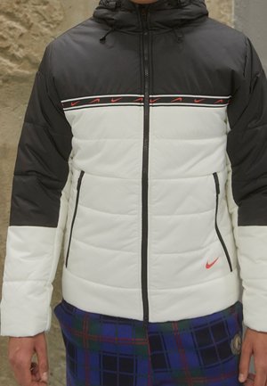 Sort og hvid dunjakke med røde Nike-logoer over brystet og tæt ved taljen, båret over blå og grøn ternede bukser.