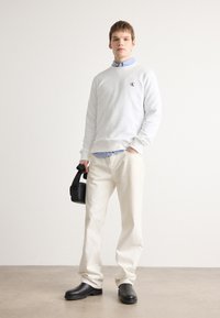 Sudadera blanca con logo bordado, camisa a rayas azul claro debajo, pantalones blanco sucio, botas negras y sosteniendo un pequeño bolso negro.