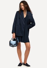 Blazer navy con doppi bottoni, indossato sopra un vestito navy. Scarpe nere senza lacci e una piccola borsa blu con dettagli in catena.