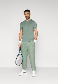 Ανδρικό αθλητικό σύνολο σε απαλό πράσινο χρώμα· κοντομάνικο πόλο με γιακά και λευκές λεπτομέρειες, ταιριαστός φόρμας παντελονιού και ρακέτα τένις.