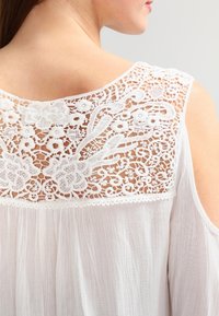 Blouse blanche avec détails en dentelle florale au dos, manches courtes, et une texture légère et transparente. L'encolure est arrondie avec des accents de bordure.