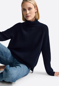 Suéter de cuello alto de punto navale con textura acanalada, corte relajado y aberturas laterales, combinado con jeans de mezclilla azul claro.