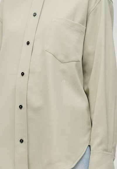 Ανοιχτό μπεζ πουκάμισο από υφασμένο ύφασμα με μυτερό γιακά, μαύρα κουμπιά, μία τσέπη και ένα μικρό λογότυπο στο πλάι.