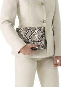 Hexagonal-patterned, snake print clutch bag in neutral tones, held against a pale cream jacket and matching pants. Flat design with flap closure.