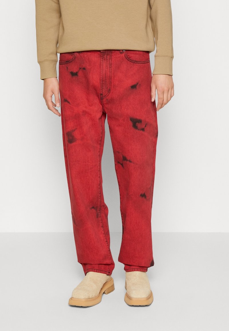 Pantaloni in denim rossi con una vestibilità rilassata, caratterizzati da un effetto tie-dye in tonalità più scure, abbinati a scarpe beige senza lacci.