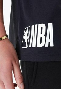 T-shirt blu navy con stampa bianca del logo NBA e grafica silhouette all'orlo, abbinato a pantaloni sportivi neri, che mostra una mano con un bracciale d'oro.