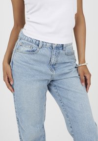 Osoba nosząca jasnoniebieskie dżinsy i białą bezrękawnik, z srebrną bransoletką na prawej nadgarstku na jednolitym tle.