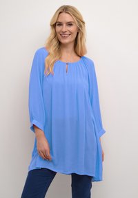 Uśmiechnięta kobieta z długimi blond włosami, nosząca luźną, jasnoniebieską bluzkę z rękawami 3/4 oraz ciemnoniebieskie spodnie, na jednolitym białym tle.