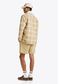 Camisa de franela beige con patrón de cuadros blancos, cuello crema, pantalones cortos caqui, zapatillas marrones y calcetines blancos con rayas rojas y azules.