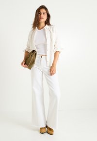 Jeans anchos blancos con dobladillos deshilachados, una camisa de botón de patrón claro y una camiseta blanca ajustada. Sosteniendo un bolso bandolera verde.