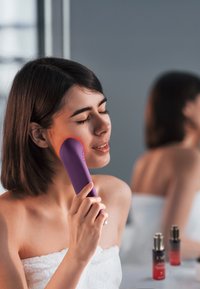 Purple facial device held against the cheek, smooth texture, ergonomic shape. Background includes two small bottles with gradient colors.