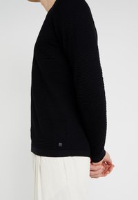 Maglione nero con texture, colletto rotondo e maniche lunghe. Presenta un orlo e polsini a coste. Piccola patch con logo vicino al fondo.