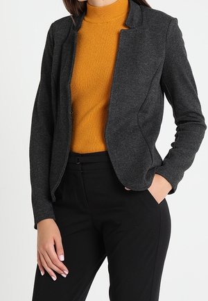 Kobieta ubrana w dopasowaną ciemnoszarą marynarkę, musztardowożółty prążkowany golf i czarne spodnie z wysokim stanem, stojąca z jedną ręką w kieszeni.