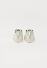 Balti odiniai sportbačiai su sidabriniais akcentais ant kulno ir kreminės spalvos juosta, pasižymintys lygiomis tekstūromis ir apvalia forma.