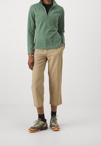 Lichtgroene fleece pullover met een half-rits kraag, beige cropped broek en meerkleurige wandelschoenen met oranje accenten en een gestructureerde zool.