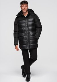 Cappotto a sacco nero con cappuccio, caratterizzato da una finitura lucida, un motivo a righe orizzontali e due tasche frontali. Indossato sopra pantaloni e scarpe neri.