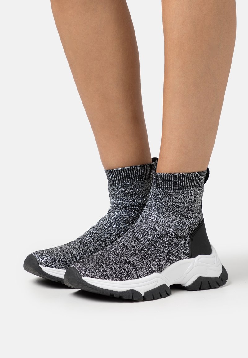 Jambes portant des baskets chaussettes tricotées noir et blanc avec des semelles blanches épaisses et des semelles extérieures en caoutchouc noir sur un fond blanc uni.