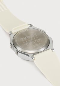 Montre Timex avec un fond en acier inoxydable, un bracelet en silicone crème, deux boutons latéraux et des détails gravés au dos.