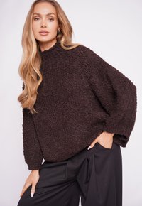 Brown textured oversized sweater with a high neck and long sleeves, paired with black pants. Soft, cozy material creates a relaxed fit.