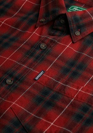 Camisa de franela a cuadros rojos y negros, con botones marrones, un patrón de cuadrícula blanca y una etiqueta azul de Superdry en el frente.
