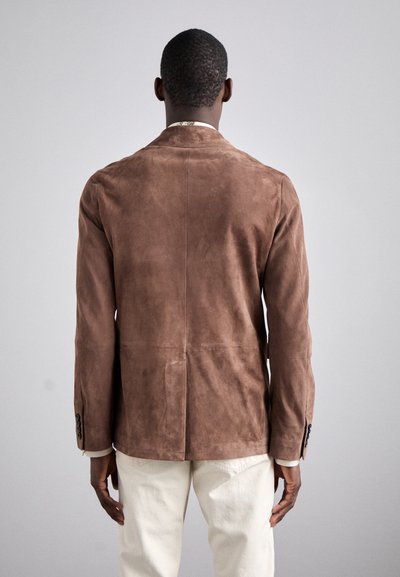 Blazer en daim marron avec une coupe ajustée, revers crantés, une fente arrière unique et des poignets boutonnés. Porté avec un pantalon de couleur claire.