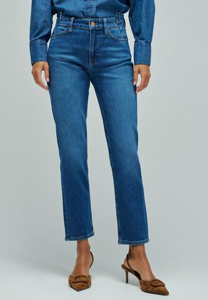 Persona con vaqueros azules de pierna recta y zapatos de tacón marrones con tira trasera, de pie frente a un fondo liso.