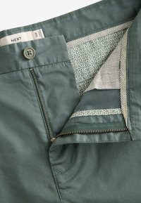Pantaloni di cotone verdi con una texture liscia, dotati di chiusura con bottone, zip e fodera interna con motivo geometrico.